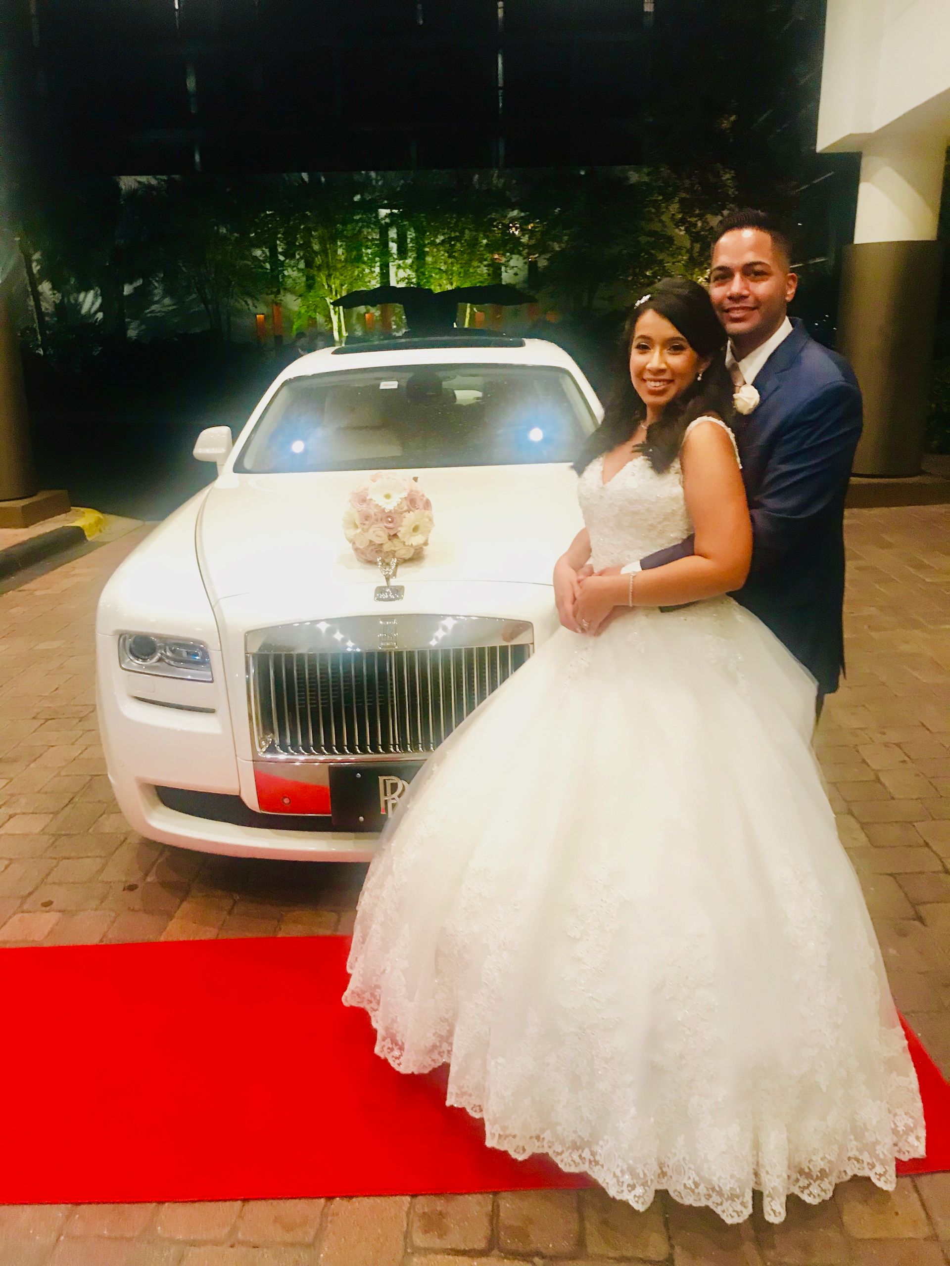 Couple poses in front of white Rolls Royce on red carpet, woman in white wedding dress, man in blue suit.Exotic Limo Orlando