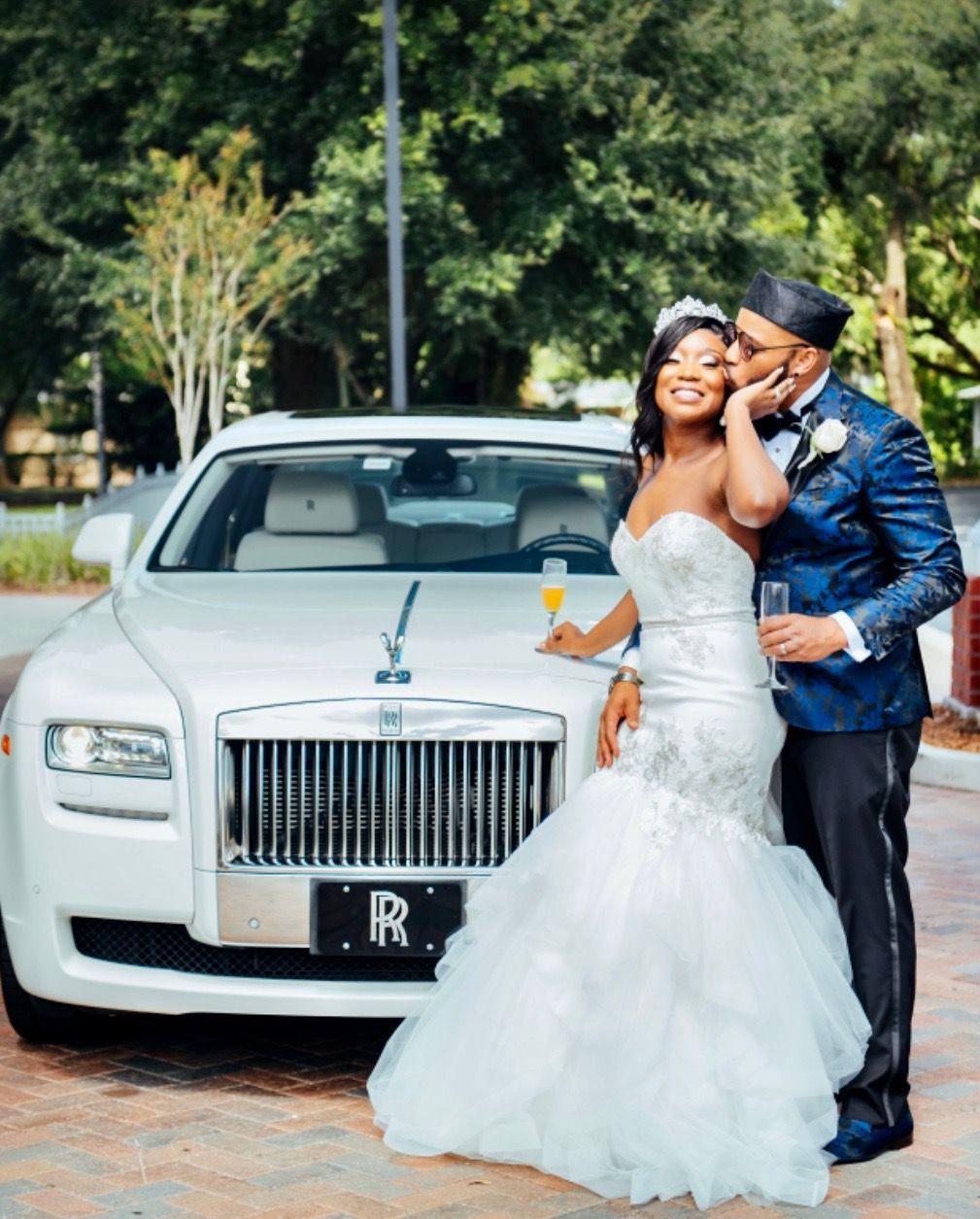 Bride and groom in wedding attire pose by a white Rolls Royce. She smiles, holding champagne; he kisses her cheek.