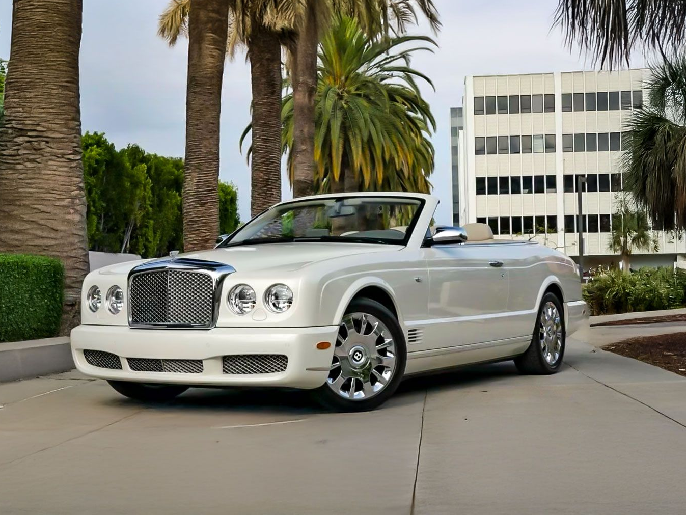 White Bentley convertible parked on a paved driveway in front of palm trees and a building.