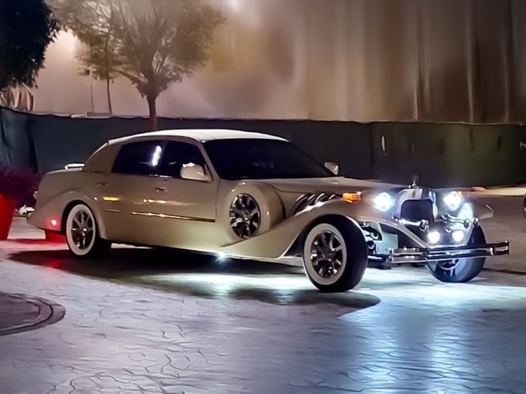 Cream-colored, vintage-style limousine with bright headlights parked at night, reflecting on wet pavement.