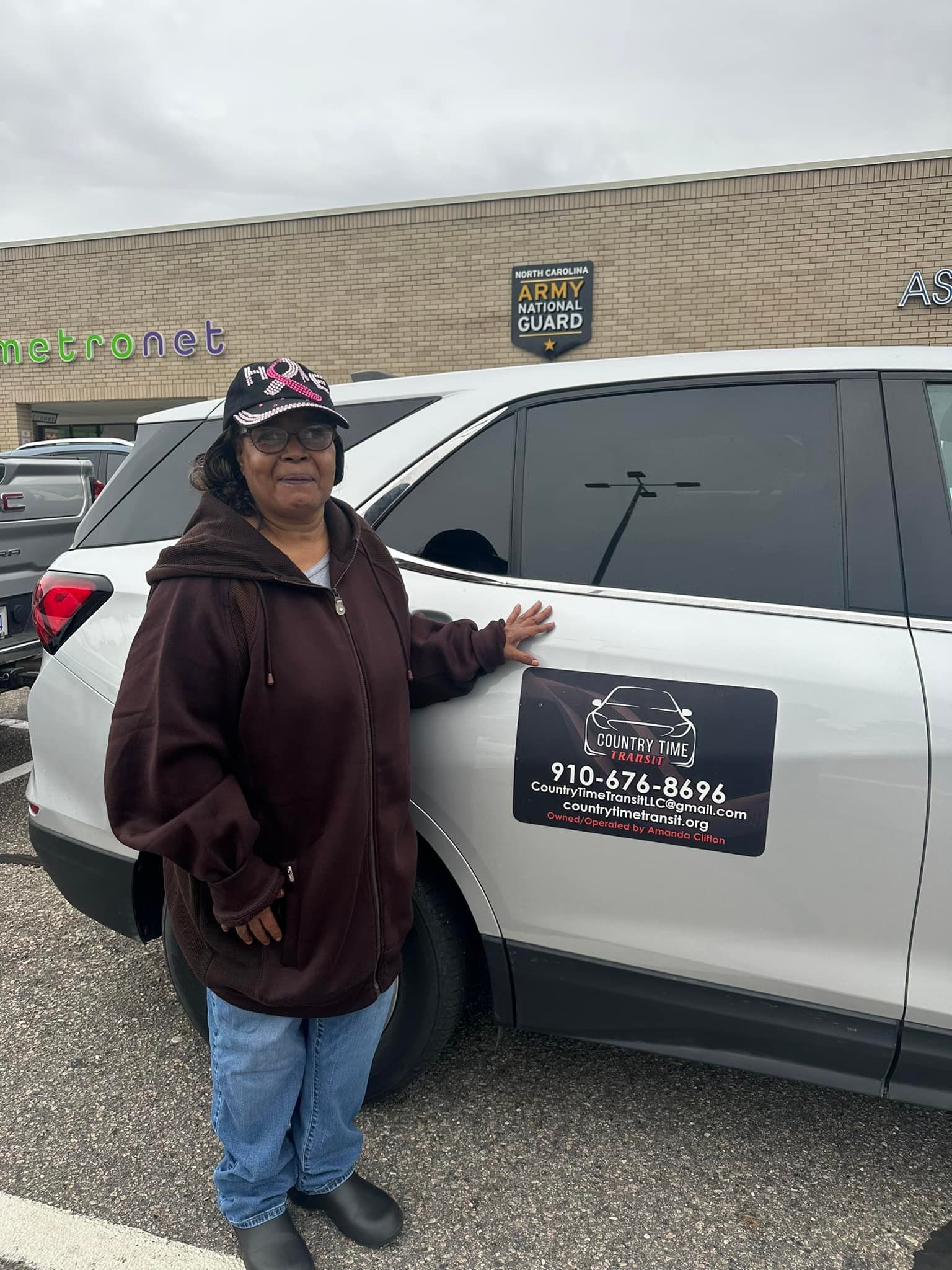 Woman in brown jacket poses by a white SUV with a sticker. A store is in the background.