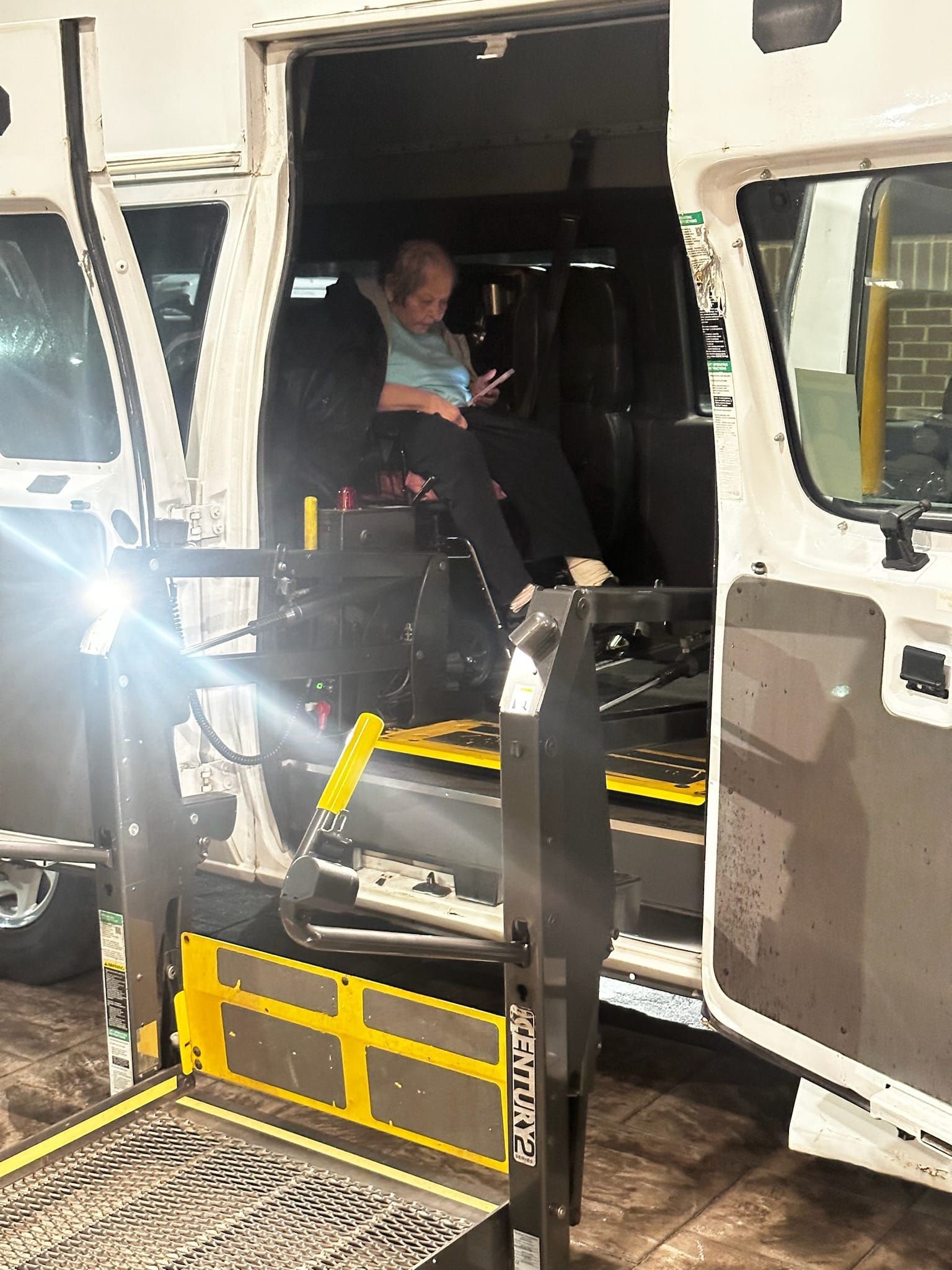 Person seated in a vehicle using a wheelchair lift. The lift is lowered, vehicle doors are open.