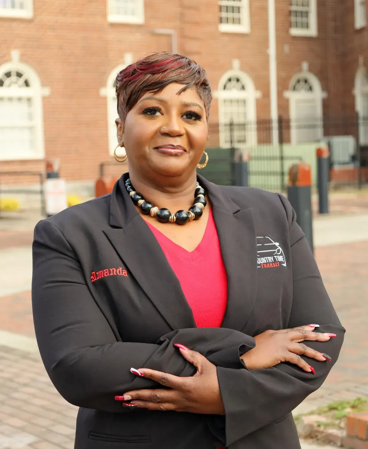 Woman in black blazer, red top, arms crossed, standing outdoors near a brick building.