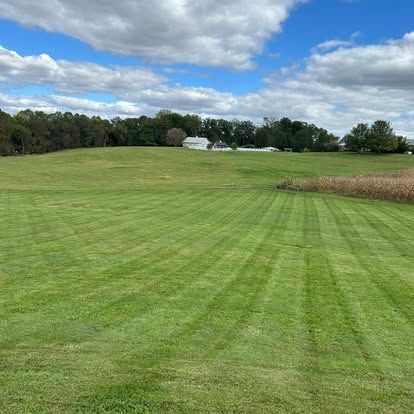 Wide green mowed field under cloudy sky, with trees and a distant farmhouse.