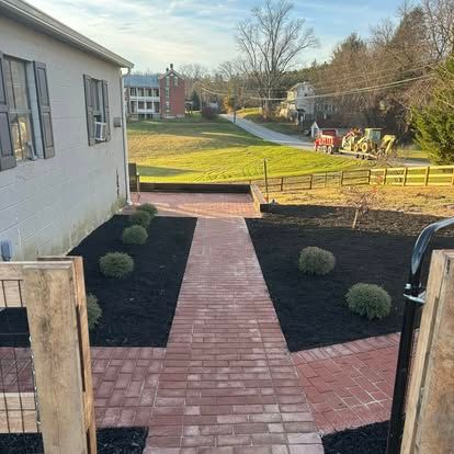 Brick walkway from a house to a grassy yard, with landscaped shrubs and a distant red tractor.