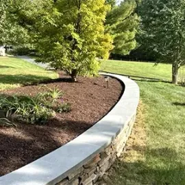 Curved stone garden wall with a concrete cap beside mulch, grass, and leafy trees in a park-like yard