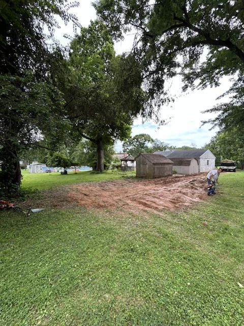 Grassy yard with large trees, a dirt patch, a small shed, and a motorcycle near a white house.