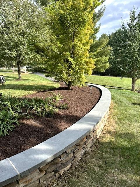 Curved stone-edged garden bed with brown mulch, green shrubs, and a sunlit tree in a grassy park.