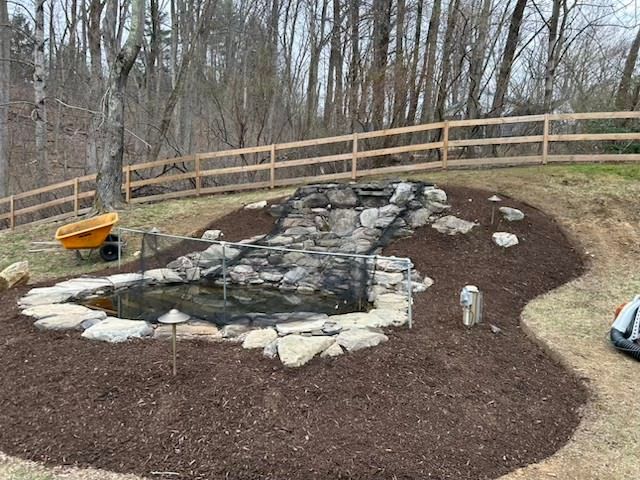Backyard landscape with a small stone pond, mulch, wooden fence, and leafless trees