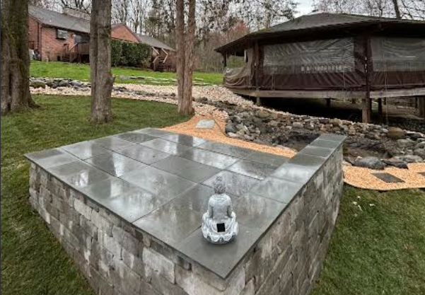 Stone memorial with a metal top and small figurine in a grassy park beside a rock-lined stream and pavilion