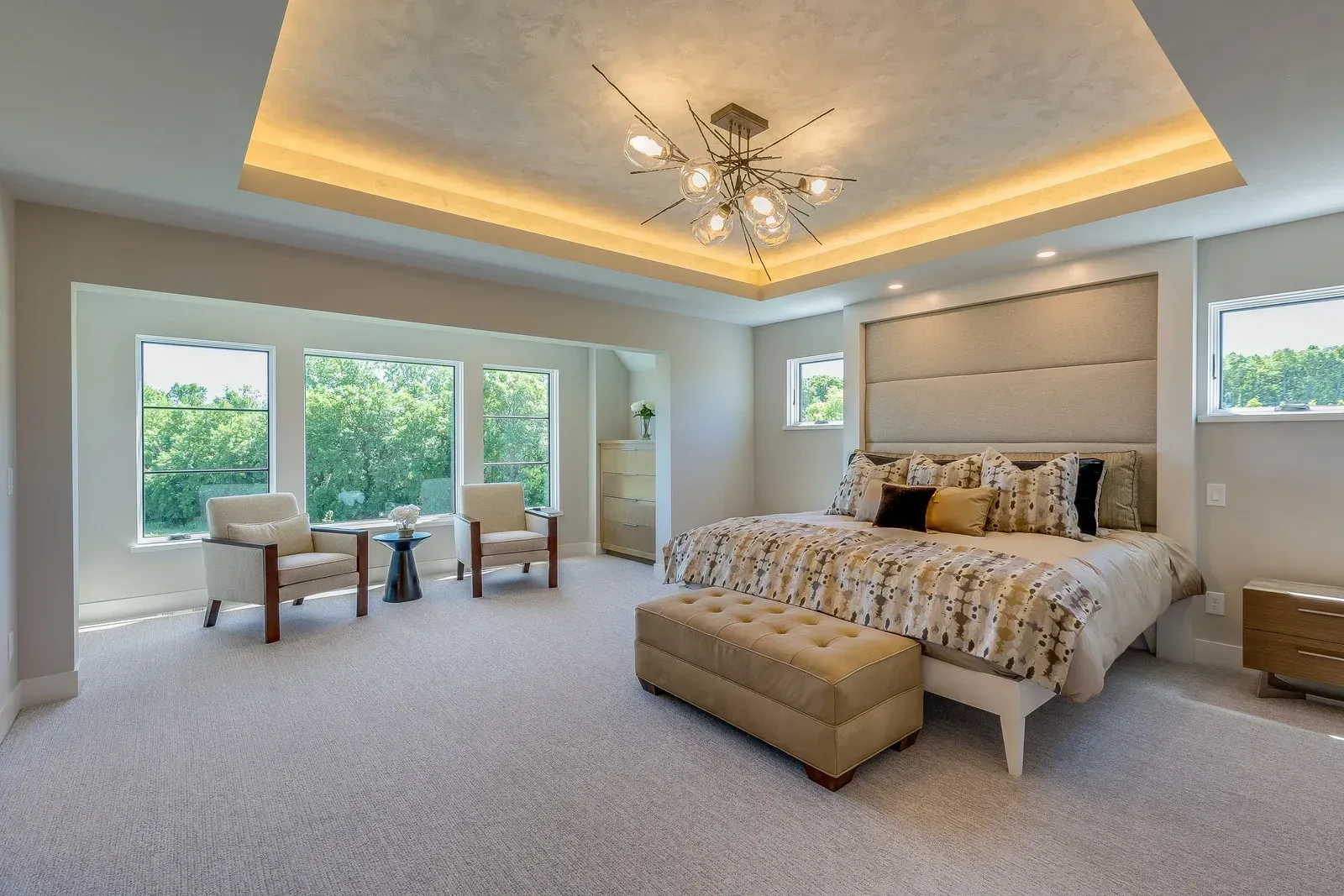 Upscale bedroom with large windows, seating area, large bed, and tray ceiling with recessed lighting.