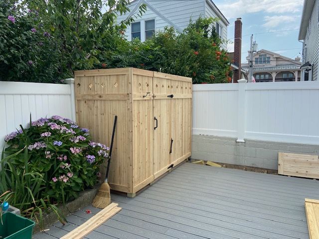 A wooden trash can is sitting on a deck next to a white fence.