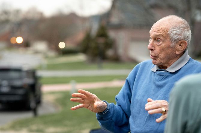 An elderly man in a blue sweater is talking to another man.