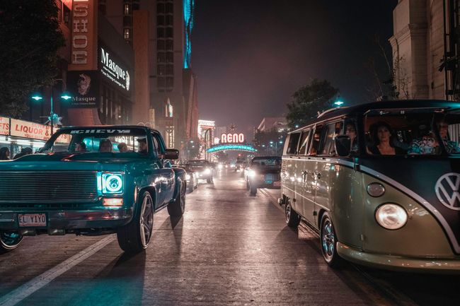 A row of cars are driving down a city street at night.