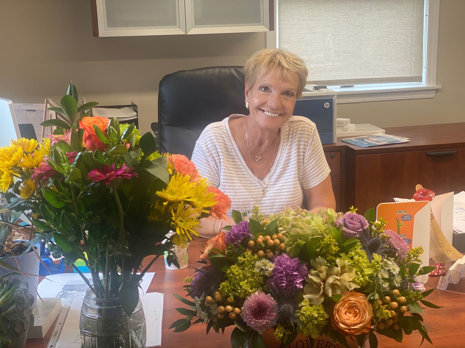 Woman with short blonde hair smiles at her desk, flanked by two colorful bouquets. She wears a white striped shirt.