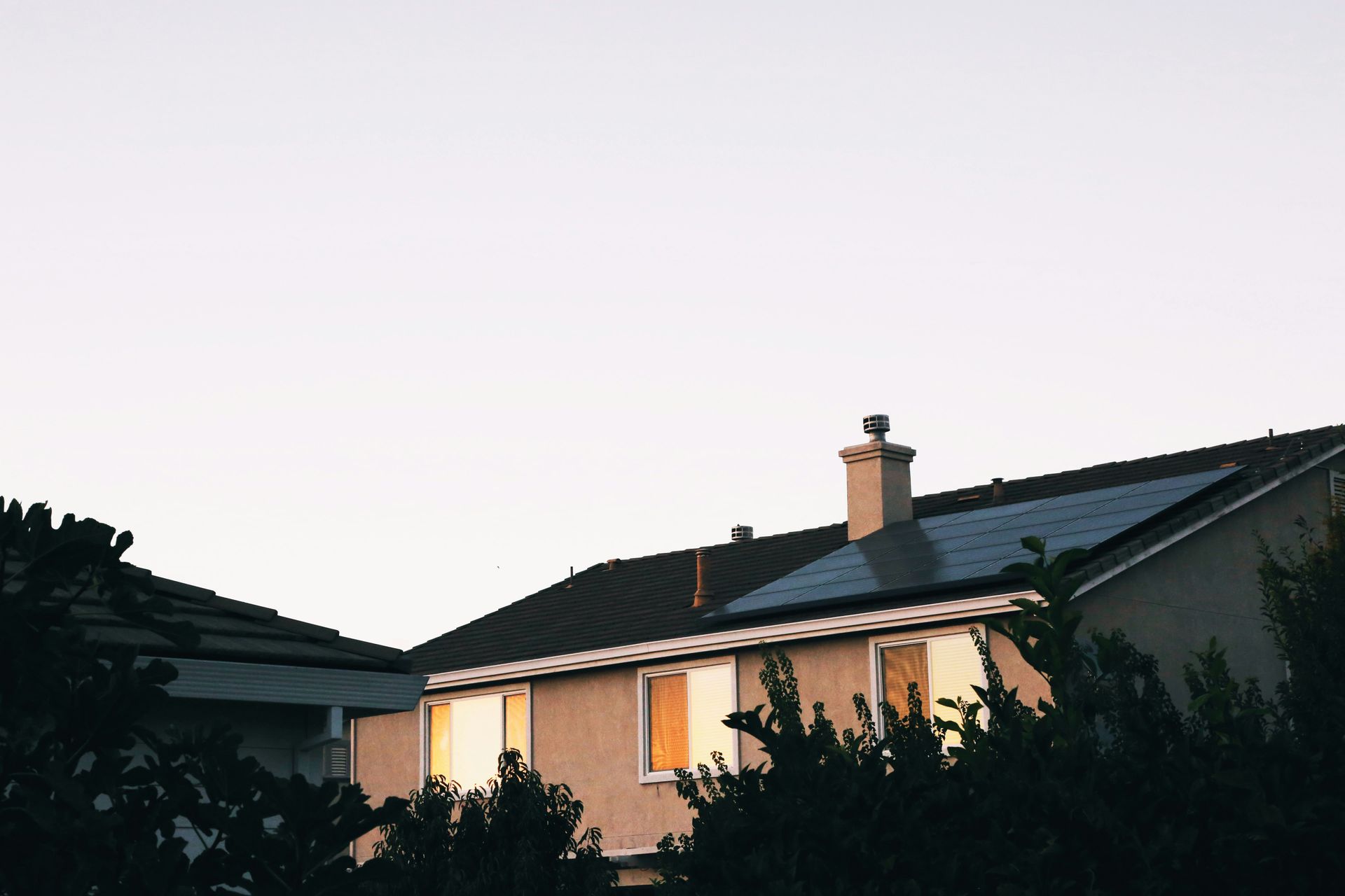 Suburban house at dusk with lit windows and dark shrubs in front