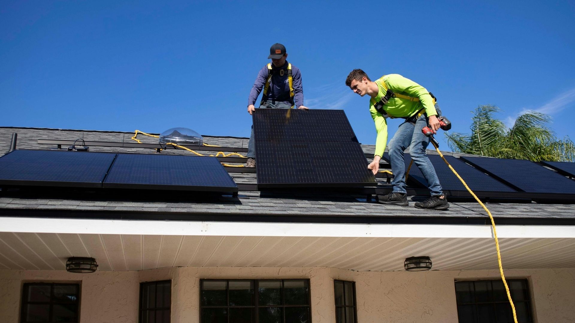 Two workers install black solar panels on a roof under a clear blue sky.