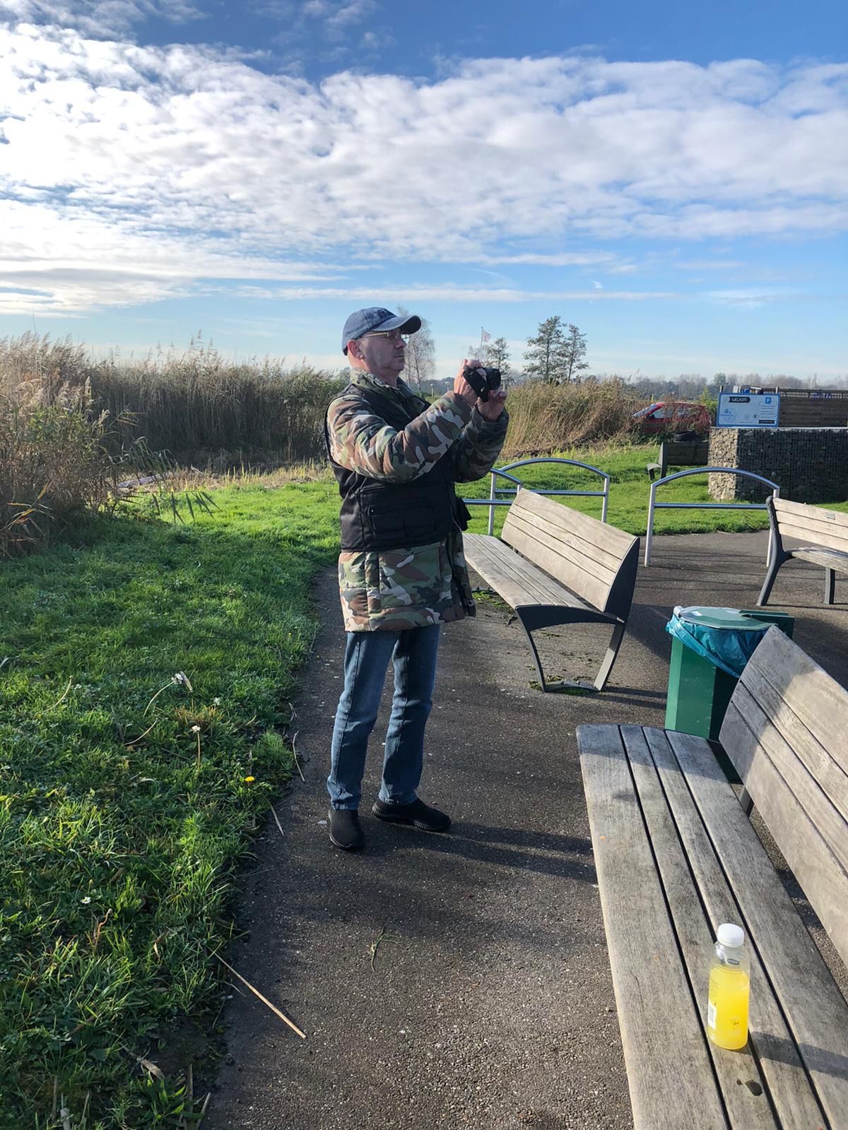 Persoon in camouflagevest maakt buiten een foto. Staat op een pad bij bankjes, hoog gras en een blauwe lucht.