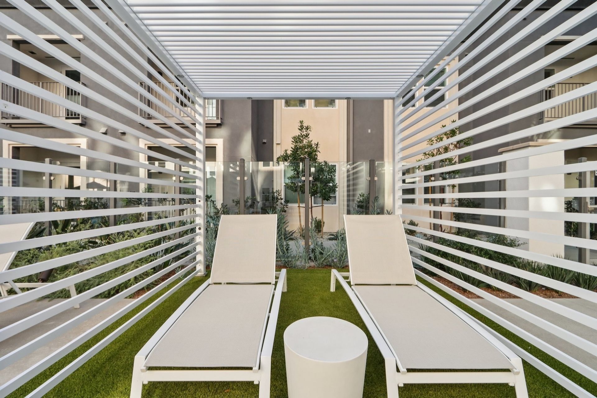 Two lounge chairs under white slat structure on artificial grass; small white side table.