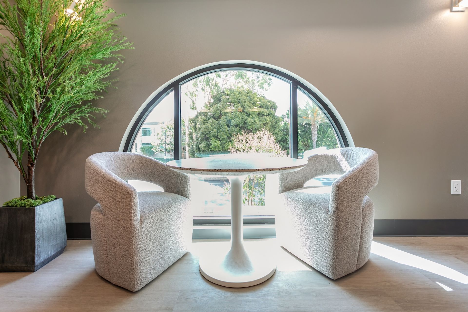 Two textured cream armchairs sit at a small white pedestal table in front of a large arched window next to a potted tree.