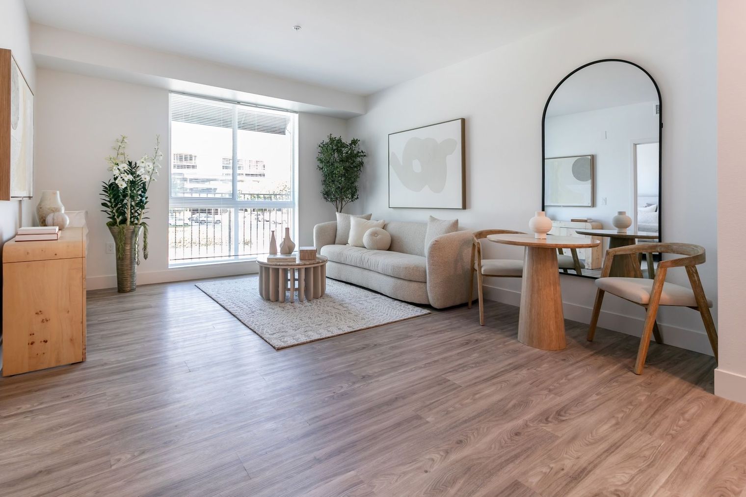 Bright modern living room with beige sofa, arched mirror, wood table, rug, plants, and large window.