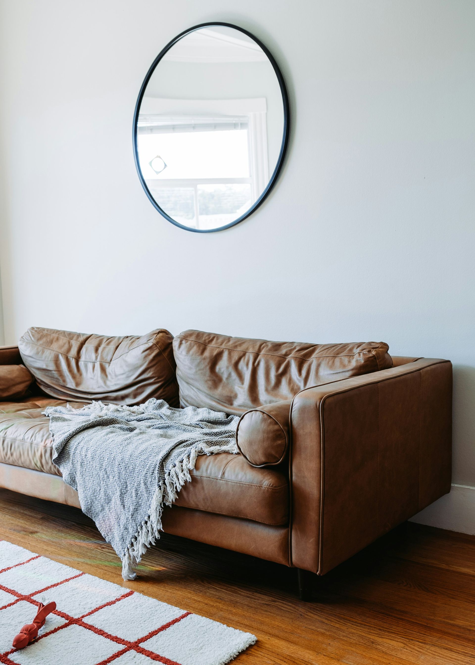 A living room with a brown leather couch and a round mirror on the wall.