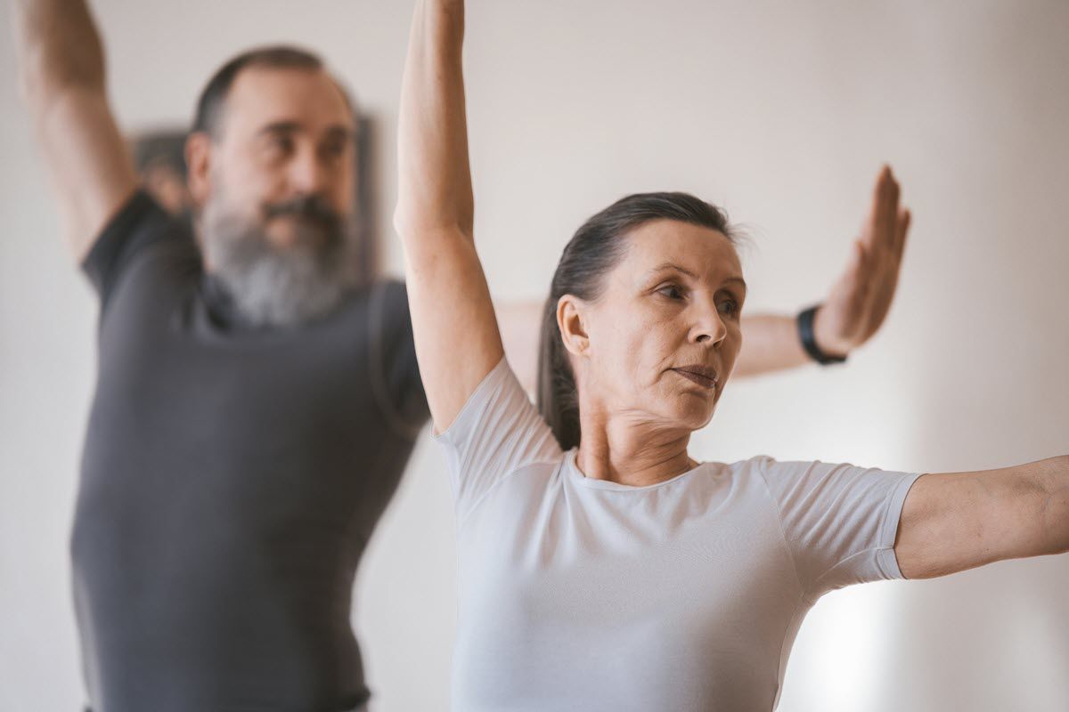 A man and a woman in athletic wear hold a yoga pose with arms extended in a bright, indoor setting.
