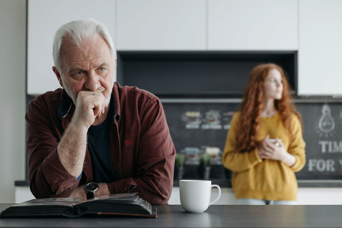 Older man thinking at a kitchen table, with a woman in the background near a chalkboard sign.