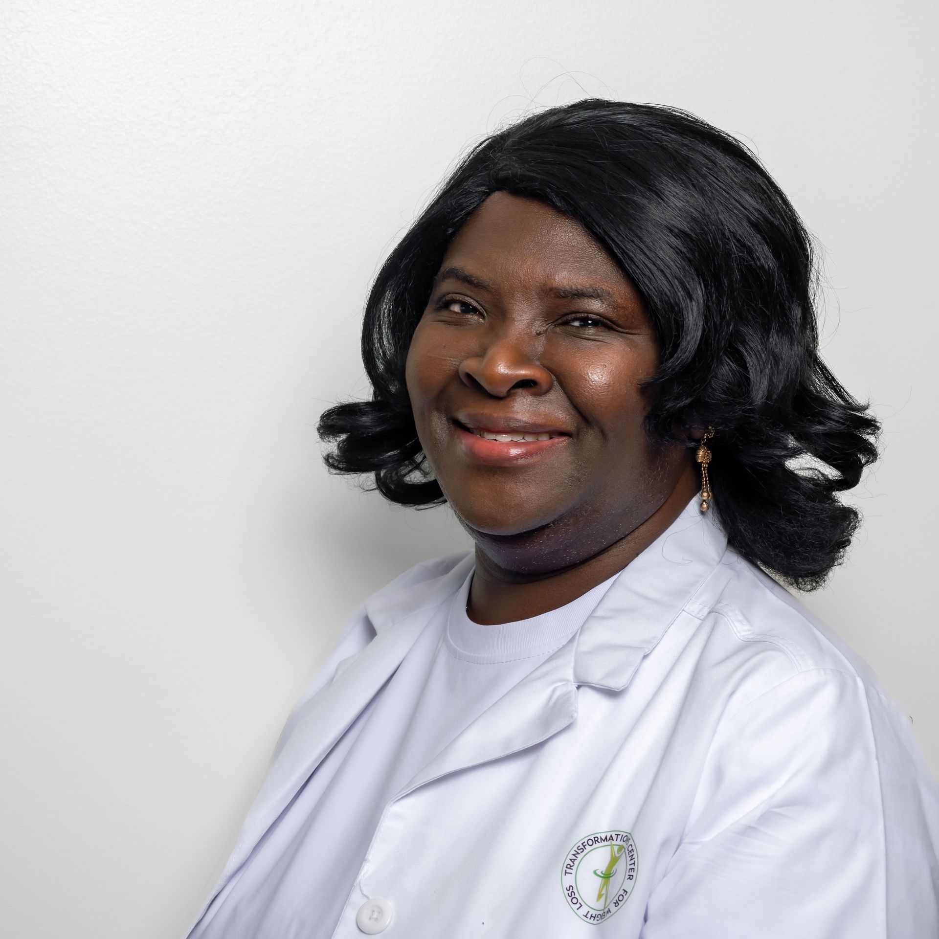 Woman in a white lab coat smiles. She has black hair and is against a white background.