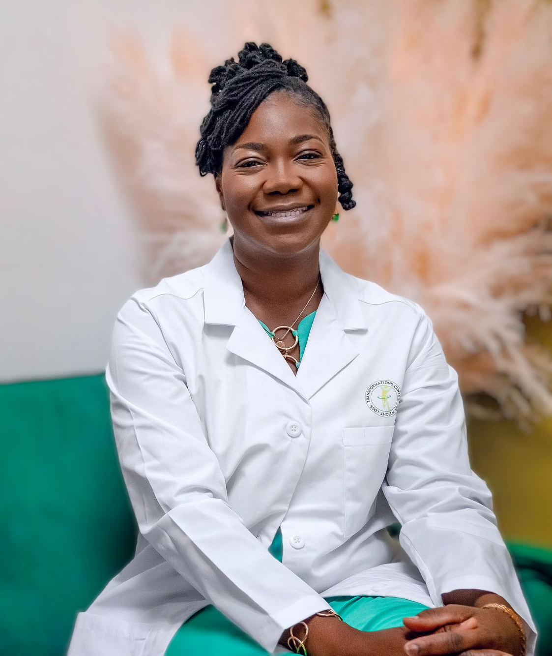 Woman in white lab coat smiles; seated, green background.