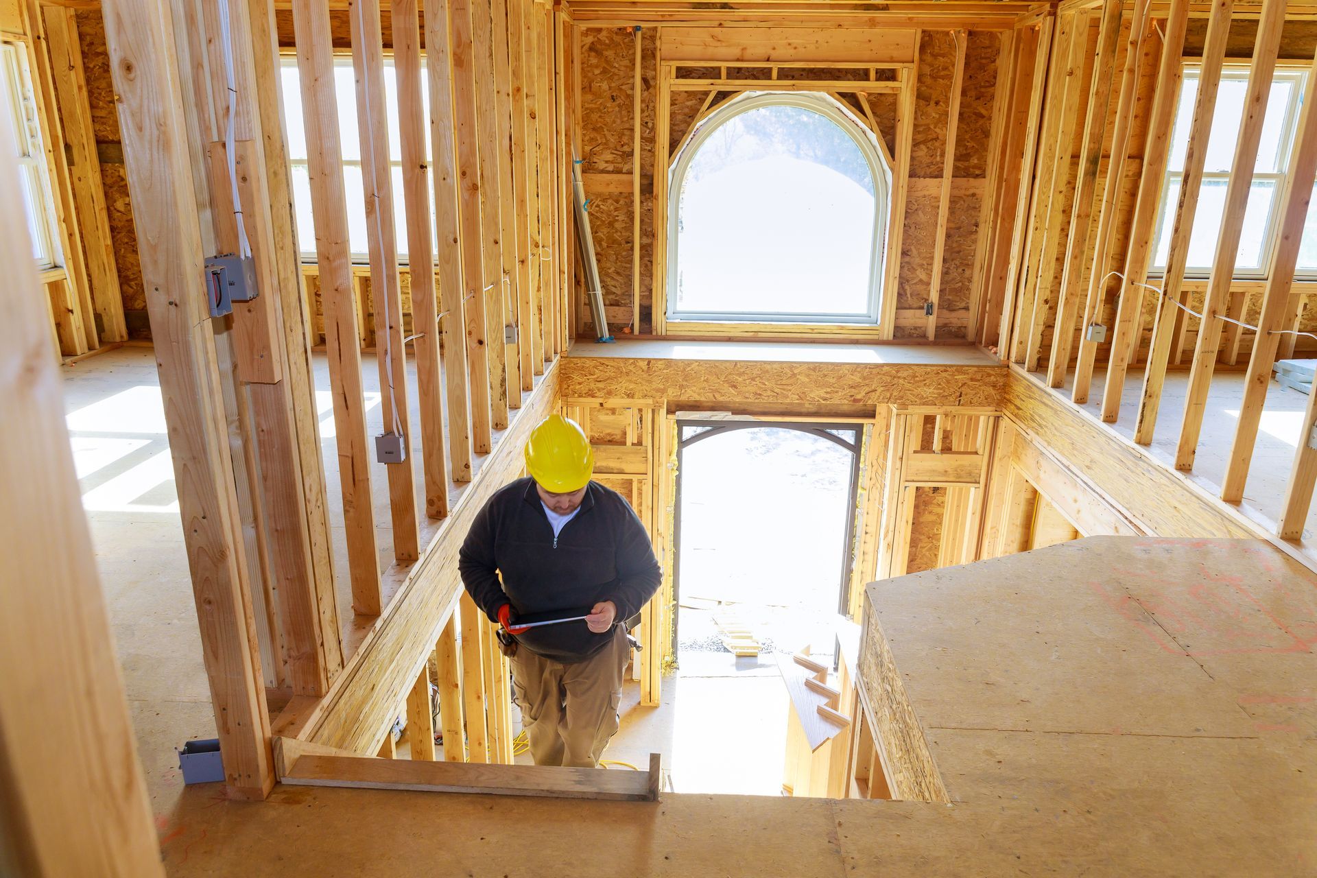 Construction worker in a partially built house, descending stairs. Yellow hard hat, light wood, bright windows.