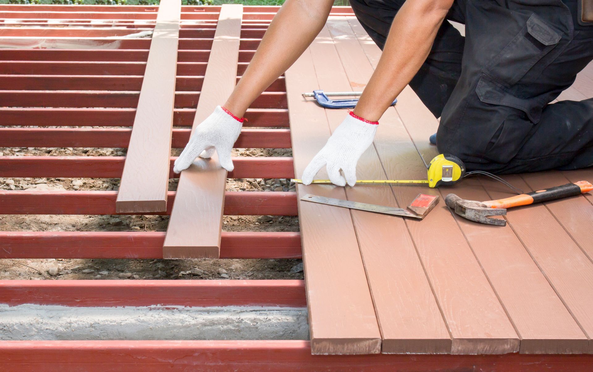 A person installing a composite deck, measuring and positioning boards over red support beams, outdoors.