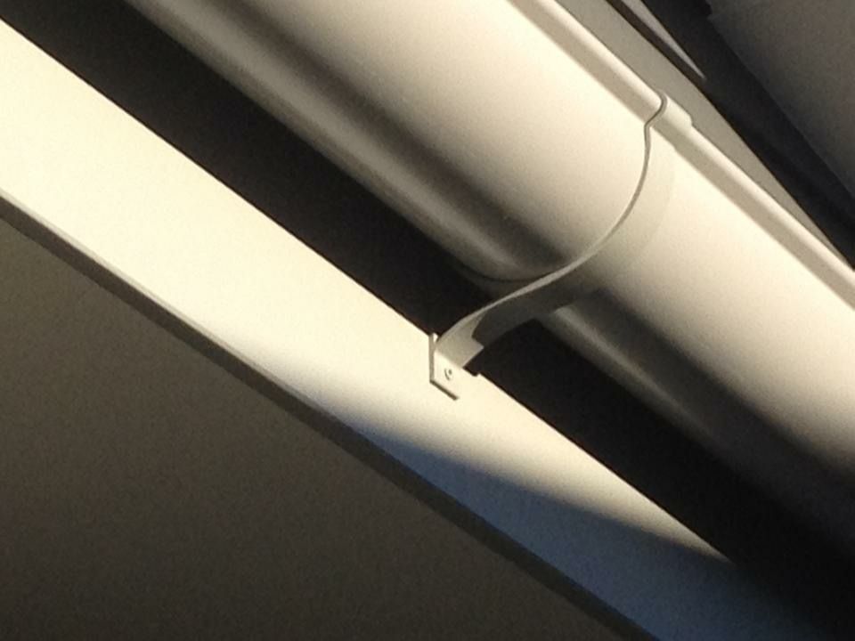 White gutter pipe held up by a matching bracket attached to a white beam. Sunlight casts shadows.