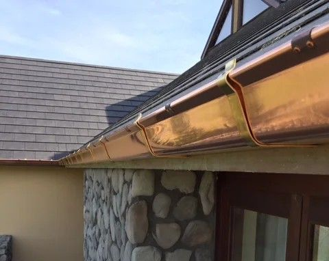 Gray rain gutter and downspout on the corner of a building. The roof and a portion of a chimney are visible.