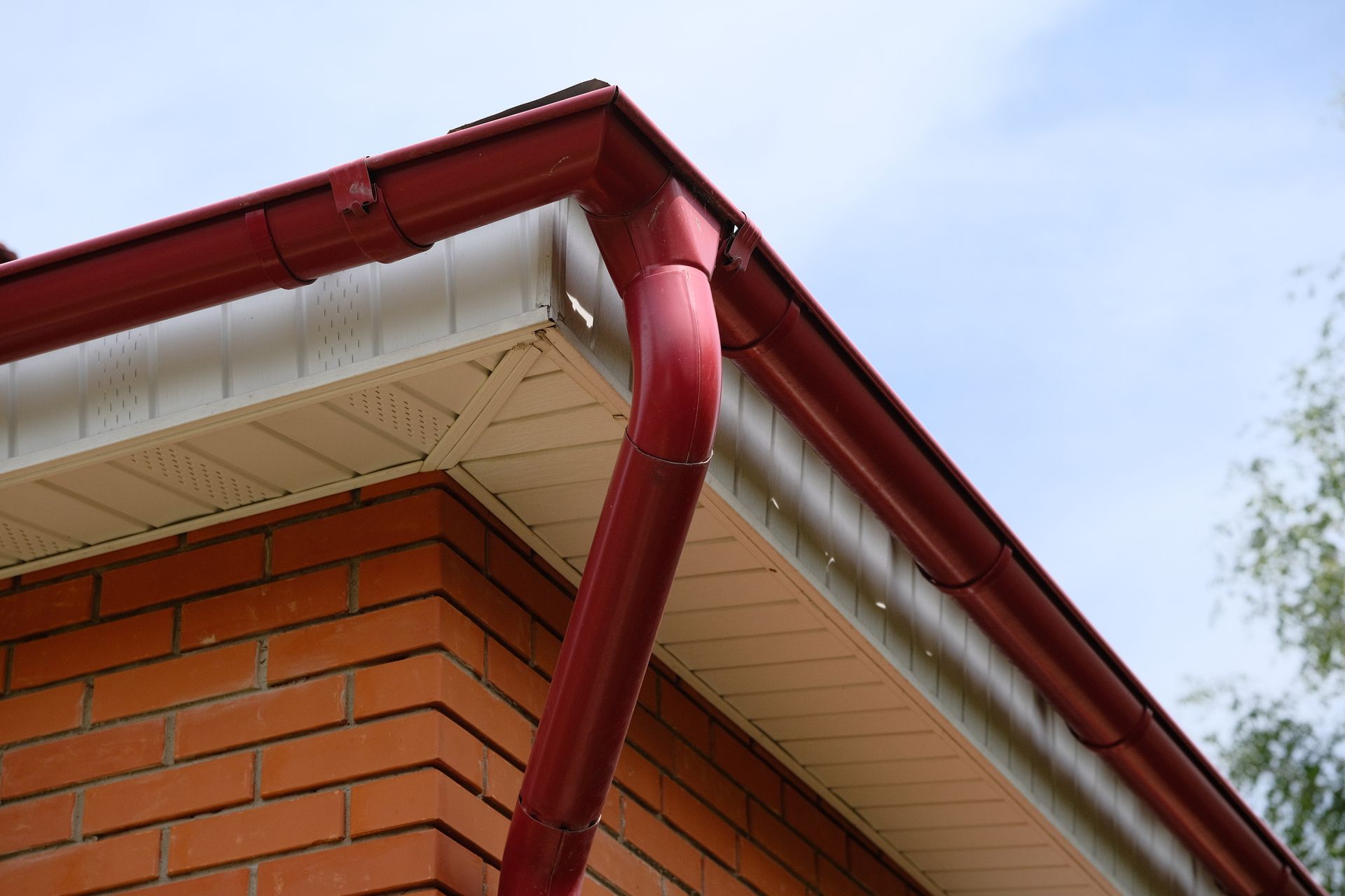 Red gutters and downspout on a brick building with white siding, against a cloudy sky.