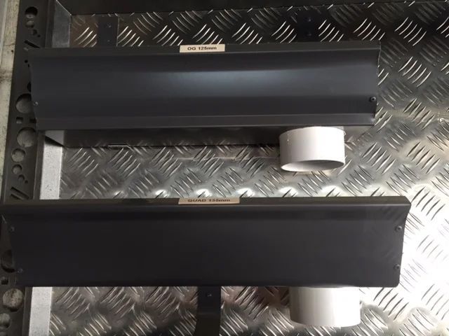 Two black rain gutters with drainage spouts are displayed against a diamond plate background.