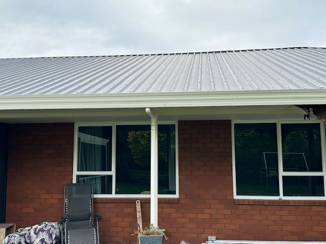 Exterior of a brick building with a corrugated metal roof, white gutters, and windows. A chair and potted plant are in the foreground.