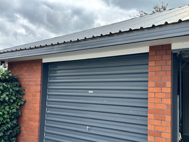 Brick garage with a gray corrugated metal door and roof. A gray gutter and trim contrast with the red brick and cloudy sky.