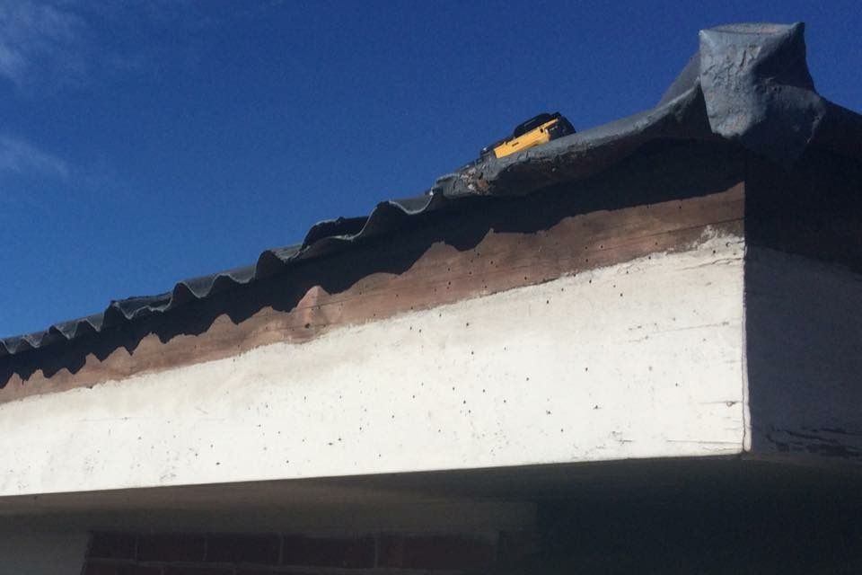 Close-up of a building's roof edge against a blue sky. The roof appears to be made of corrugated dark material over a white concrete structure.