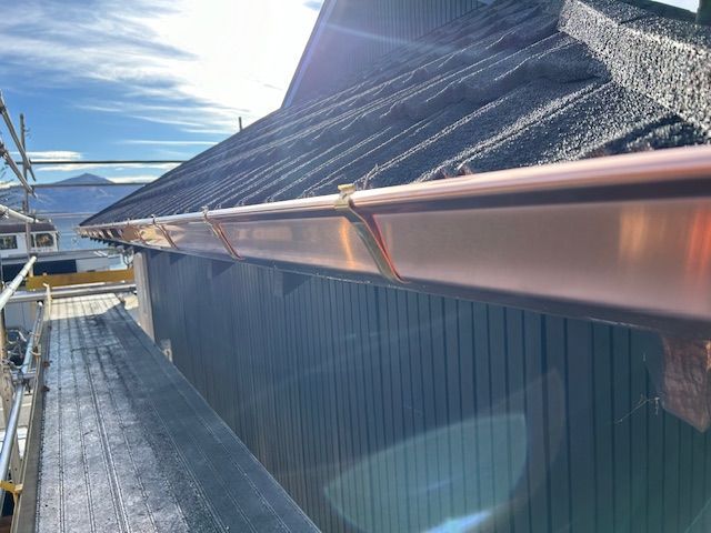 Copper gutters installed on a dark-roofed building; scaffolding and a mountainous background visible.
