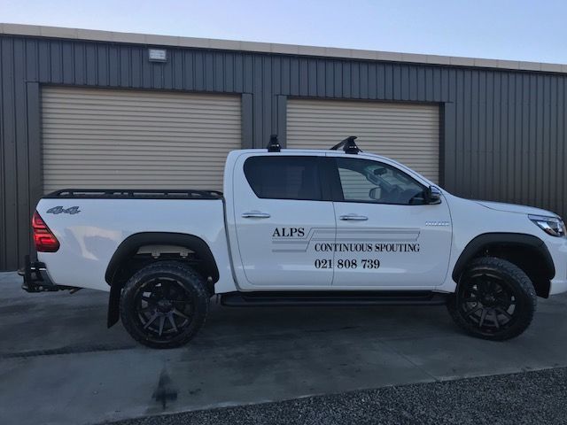 White pickup truck with black wheels, rack, and company logo parked in front of a gray building.