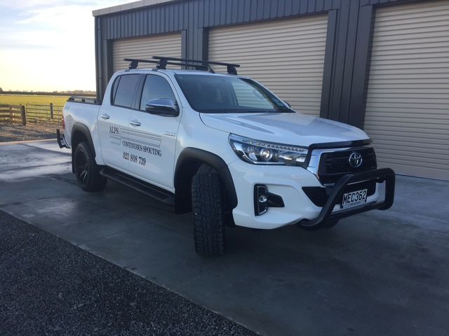 White Toyota Hilux pickup truck with a black bull bar parked on a concrete surface in front of a building with roller doors.