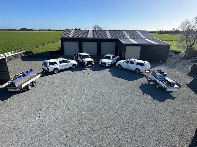 White trucks and boat trailers parked in front of a large shed on a gravel surface in a rural setting. Blue sky.