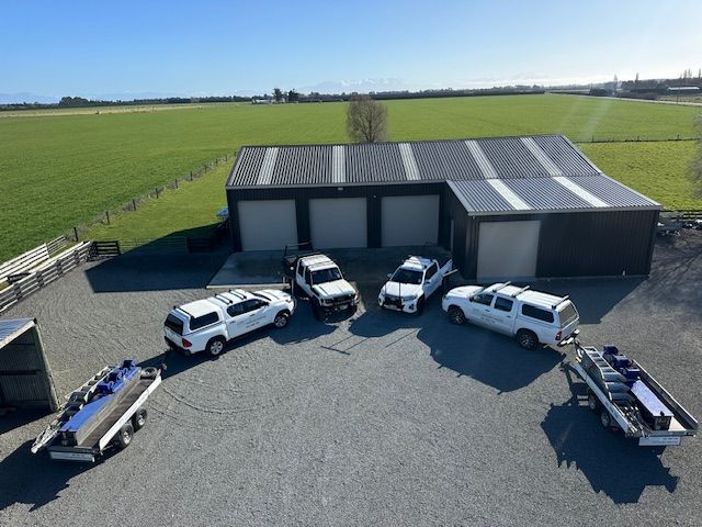 Five white vehicles and two trailers parked outside a dark garage in a grassy field.