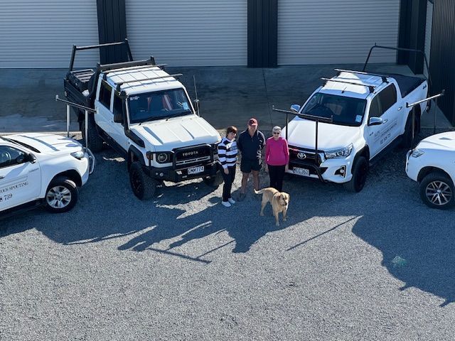 Three people and a dog stand in front of four white trucks. The trucks are parked on gravel with a gray building in the background.
