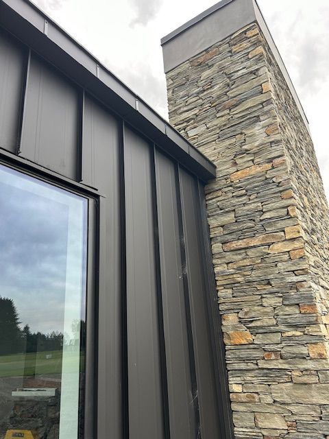 Black metal siding and trim on a modern building next to a stacked stone chimney. A window reflects an overcast sky.