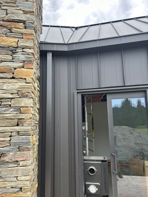Exterior view of a modern building with gray metal siding, a stone chimney, and a glass door. A ladder is visible inside the door.