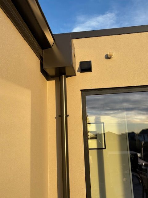 Dark brown gutter system on the corner of a light-colored building. Vertical downspout runs from the gutter towards the ground.