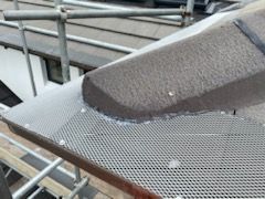 Close-up of a gray metal gutter guard with a dark, textured roof component. The gutter guard is mounted on a roof, with scaffolding in the background.