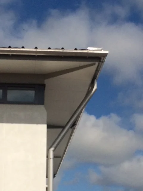 Close-up of the corner of a white building with grey trim, gutters, and downspout against a blue sky with clouds.